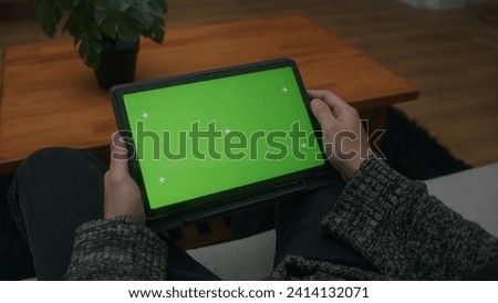 Young man scrolling and tapping center on tablet with green screen mock up display. Male sitting on sofa, relaxing at home. Close-up over the shoulder shot