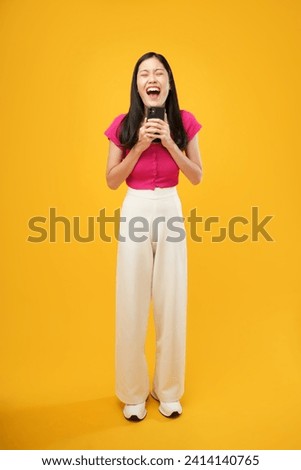 Full body photo of young Asian woman holding phone with cheerful face isolated on yellow background