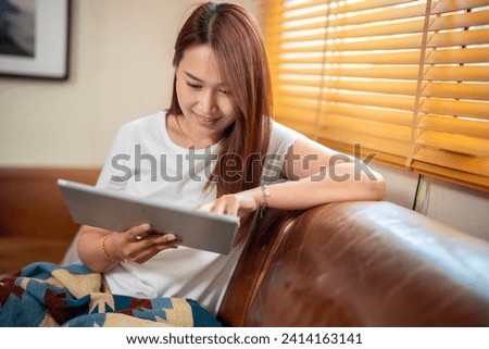 Happy pretty young woman in casual outfit sitting on sofa at home and using tablet while browsing internet