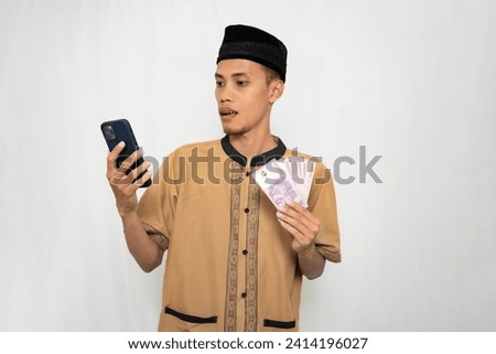 Asian Muslim man wearing Muslim clothes is shocked looking at the smartphone screen while carrying money in his other hand. Isolated white background.