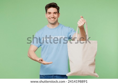 Young man wears blue t-shirt white title volunteer hold in hand point on eco cotton textile shopper isolated on plain pastel green background. Voluntary free work assistance help charity grace concept