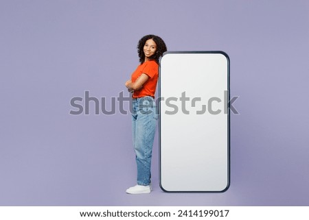 Full body little kid teen girl of African American ethnicity wear orange t-shirt stand near big huge blank screen area mobile cell phone isolated on plain purple background Childhood lifestyle concept