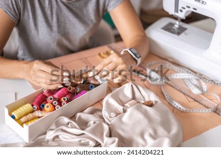 Woman dressmaker hands choosing thread color for sewing beige fabric clothes at workshop studio closeup. Female tailor fashion designer art work modist dressmaking professional occupation top view Royalty-Free Stock Photo #2414204221