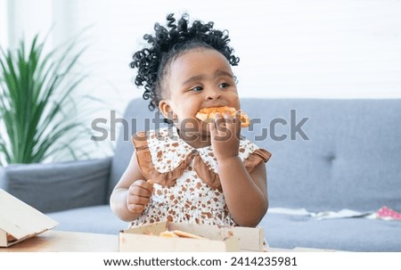 Portrait of Cute African child eating pizza at home party. Kid enjoy and having fun with tasty lunch meal. Happy little girl with yummy face holding eating pizza by her hand. Face stained with sauce