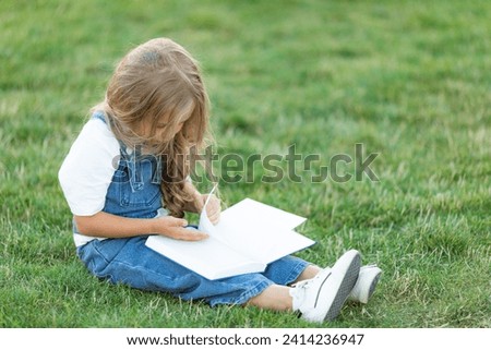 Cute little girl sitting on the grass and reading a book on summer day. Children education concept.