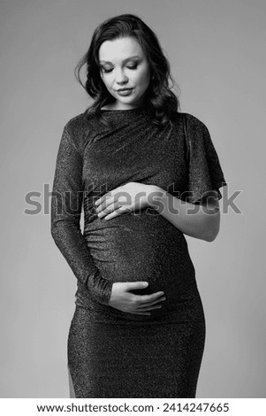 Black and white portrait of young pretty pregnant woman on gray studio background. Female in grey sequin dress with hands near pregnant belly. Royalty-Free Stock Photo #2414247665