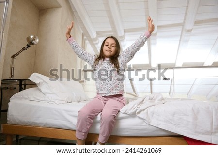 Caucasian beautiful little girl waking up in the morning, stretching her arms upwards, smiling cutely, looking at camera, sitting on the comfortable double bed with white bedsheets in her bedchamber Royalty-Free Stock Photo #2414261069