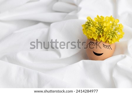 Easter egg with cute face and wreath of yellow flowers on white fabric background. Happy Easter concept