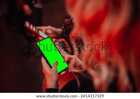 person holding a smartphone with a green screen, likely for chroma key compositing, with a blurred background.