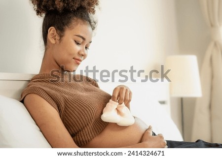 Joyful multiracial pregnant woman looking at her stomach expecting for childbirth. Young afro hair expectant mother relaxing on bed at home. Pregnancy and motherhood feminine anticipation lifestyle. Royalty-Free Stock Photo #2414324671