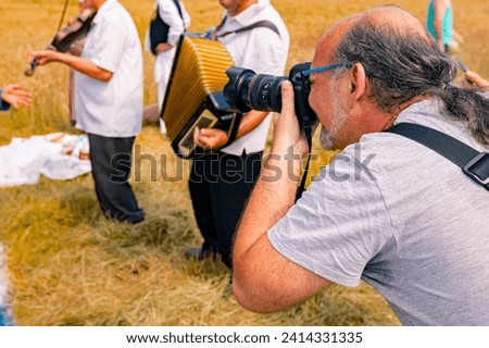 Photographer is holding photo camera, he taking pictures, looking trough camera on musicians as they play music before start reaping wheat manually in a traditional rural way.
