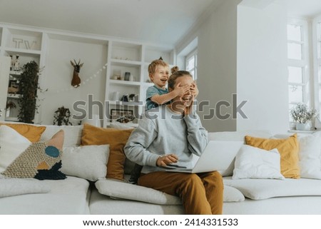 Smiling boy covering mother eyes while standing on sofa at home Royalty-Free Stock Photo #2414331533
