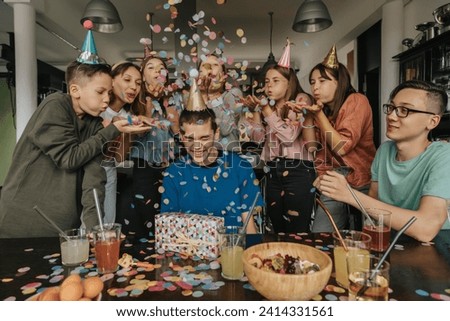 Friends throwing confetti on birthday boy sitting with gift at dining table