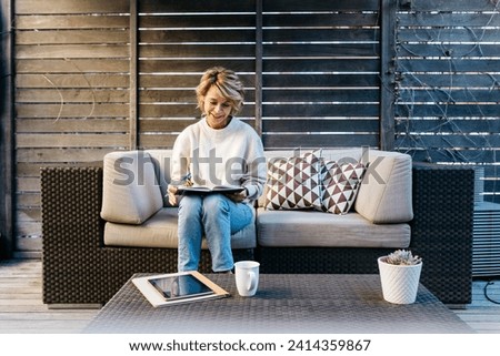 Smiling woman writing in diary while sitting on sofa at rooftop garden Royalty-Free Stock Photo #2414359867