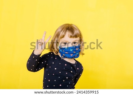 Portrait of cute blond girl wearing face mask and gesturing peace sign against yellow background