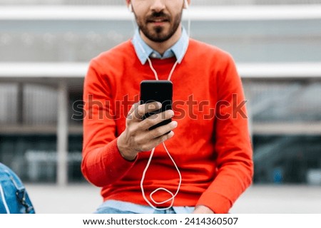 Casual businessman sitting on a bench in the city- using his smartohone and earphones Royalty-Free Stock Photo #2414360507