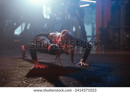 An athletic woman showcases flexibility during a push-up, with her foot extended over her shoulder, under dramatic gym lighting Royalty-Free Stock Photo #2414371023