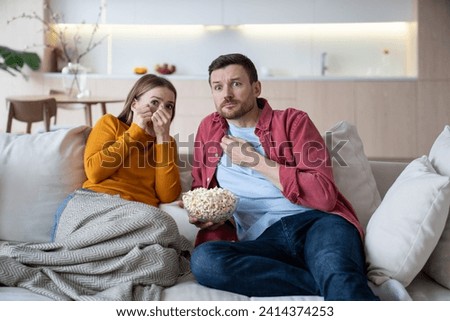 Married couple spending weekend at home, watching thriller, action film, eating popcorn. Frightened wife ready to close eyes of fearful, scary scene, intrigued husband looking with interested glance Royalty-Free Stock Photo #2414374253