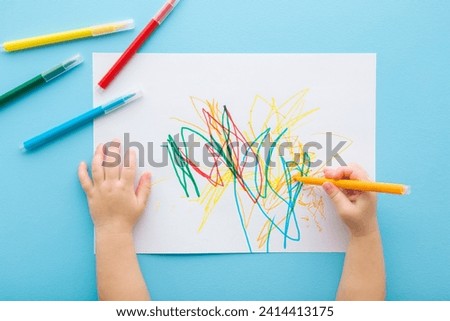 Baby boy hand holding marker and drawing first colorful scratches lines on white paper. Light blue table background. Pastel color. Closeup. Toddler development. Learning painting. Point of view shot.