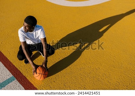 Thoughtful young man holding basketball crouching on sports court during sunny day