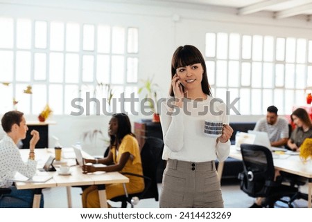 Portrait of smiling young woman on cell phone in office with colleagues in background