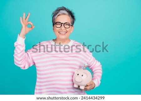 Photo of cool funky lady wear striped sweater showing okey gesture holding money box empty space isolated turquoise color background