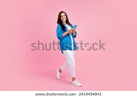 Full size photo of clever girl dressed blue shirt white trousers hold smartphone walk empty space isolated on pink color background