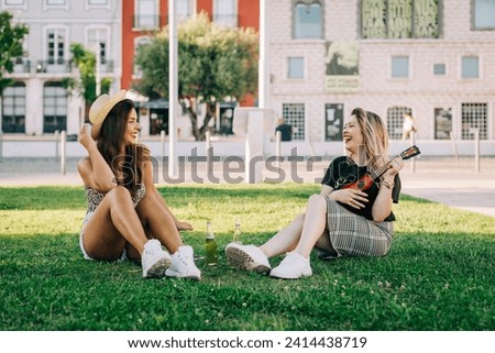 Cheerful woman playing ukulele while sitting with female friend on grassy land Royalty-Free Stock Photo #2414438719
