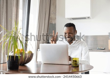 Portrait of happy man sitting at kitchen table during video chat showing victory sign
