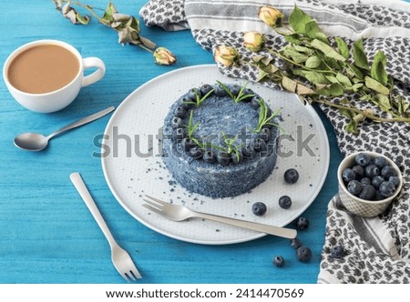 Table with Blue Blueberry Velvet cake. Cup of coffee with milk, small bowl with blueberries, scarf and dry flowers, roses.