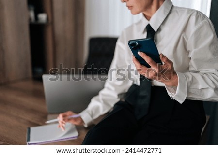 Cropped shot of successful business woman sitting on table, writing on notebook, using smartphone and working at office. Closeup female entrepreneur holding phone in hand, taking down notes on paper