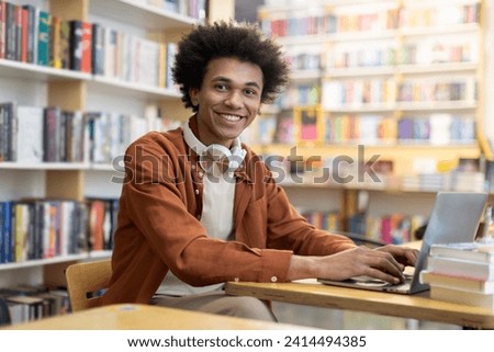 African American student guy, with headphones on neck, using laptop in college library, smiling at camera, studying online