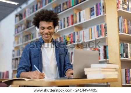 Black student guy wearing headphones and taking notes in front of laptop in university library. Dedicated study session blending technology and traditional note-taking Royalty-Free Stock Photo #2414494405
