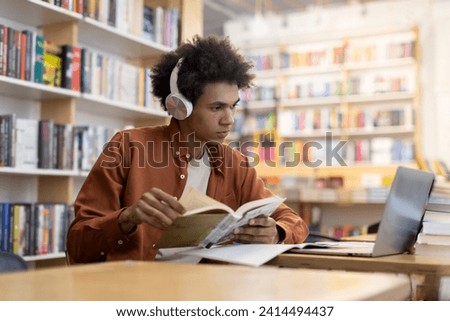 Focused black student guy in headphones looking at laptop screen and holding copybook, studying in university library, scene of concentrated focus and blended learning methods