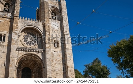 Facade of a historic cathedral with a detailed rose window and clock tower, framed against a clear blue sky with overhead tram cables, in a European city during spring.
