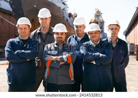 Shipyard workers at float dock wearing hard hats, stand arms-crossed. Male repair team ready for maintenance work on vessel. Maritime industry, professional ship fix group, outdoor labor day scene. Royalty-Free Stock Photo #2414532813