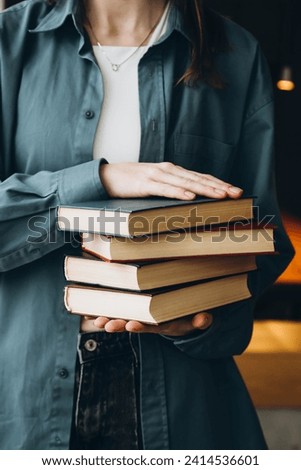 A woman in a blue shirt holds a stack of books on a dark library background. Female student in college library with books. Education, learning and reading concept.