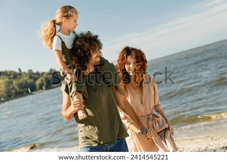 Close up shot of caucasian family carrying daughter on his shoulders on vacation on the beach. Parents holding their little kid piggyback on weekend walking along lake river