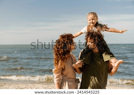 Happy family walking on the beach at sunny day. Parents looking on each other. Spouses holding their little kid piggyback on weekend walking along lake river Royalty-Free Stock Photo #2414545225