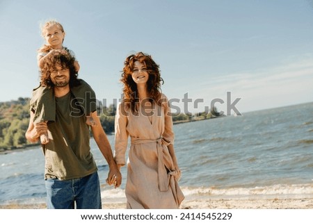 Caucasian family of mother and father looking at camera carrying daughter on his shoulders on vacation on the beach. Parents holding their little kid piggyback on weekend walking along lake river