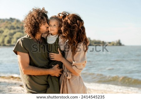 Image of young happy family kissing their daughter outdoors at the beach. Spouses father and mother parents holding their little kid child on weekend walking along lake river Royalty-Free Stock Photo #2414545235