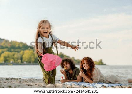 Portrait of happy little girl playing with butterfly net with parents having fun on backgroung on the beach. Active weekend with family together outdoors Royalty-Free Stock Photo #2414545281
