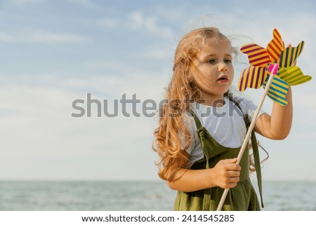 Cute little girl playing with windmill on the sunny beach. Active caucasian small daughter kid child spending time outdoors along river lake sea Royalty-Free Stock Photo #2414545285
