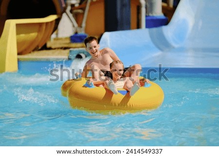 Boy and girl have fun on water slide in outdoor aquapark. Little children floating on yellow inflatable raft. Royalty-Free Stock Photo #2414549337