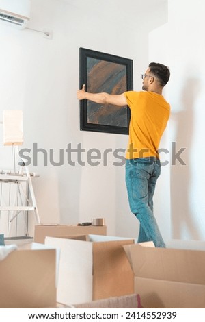 Young man decorating and putting up a framed piece of art on the wall in new home. Man in causal clothes hanging a painting on a wall. Male interior designer at work.