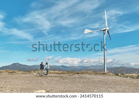 Green energy concept. Couple with dog staring the wind turbine for generating electricity in wind farm or wind park while walking near mountain top industrial wind power plant.