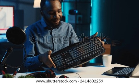 Content creator in dimly lit living room uses camera to film keyboard, mouse and headphones review for online streaming platforms. Media star hosts internet show, unboxing wired gaming peripherals