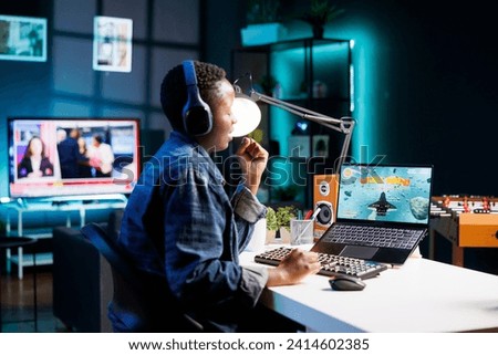 African American female gamer competes in an online video game, using laptop and wireless headset in her apartment. She aims to be the ultimate winner, fully immersed in gaming experience.
