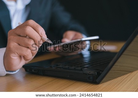 Close-up of a businessman working with a notebook computer and using a pen to take notes and using a mobile phone, smartphone, chatting, working in the office.