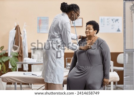 Medium long shot of mature African American woman sitting on examination table in modern clinic while female doctor listening to her heartbeat Royalty-Free Stock Photo #2414658749